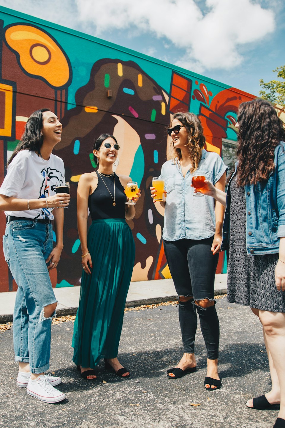Four friends laughing and holding drinks by a donut mural.